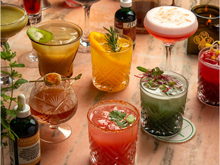 A table spread with cocktails garnished with various fruit, herbs, and tinctures