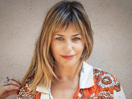 Portrait of a woman with blonde hair and bangs, in a white and orange shirt